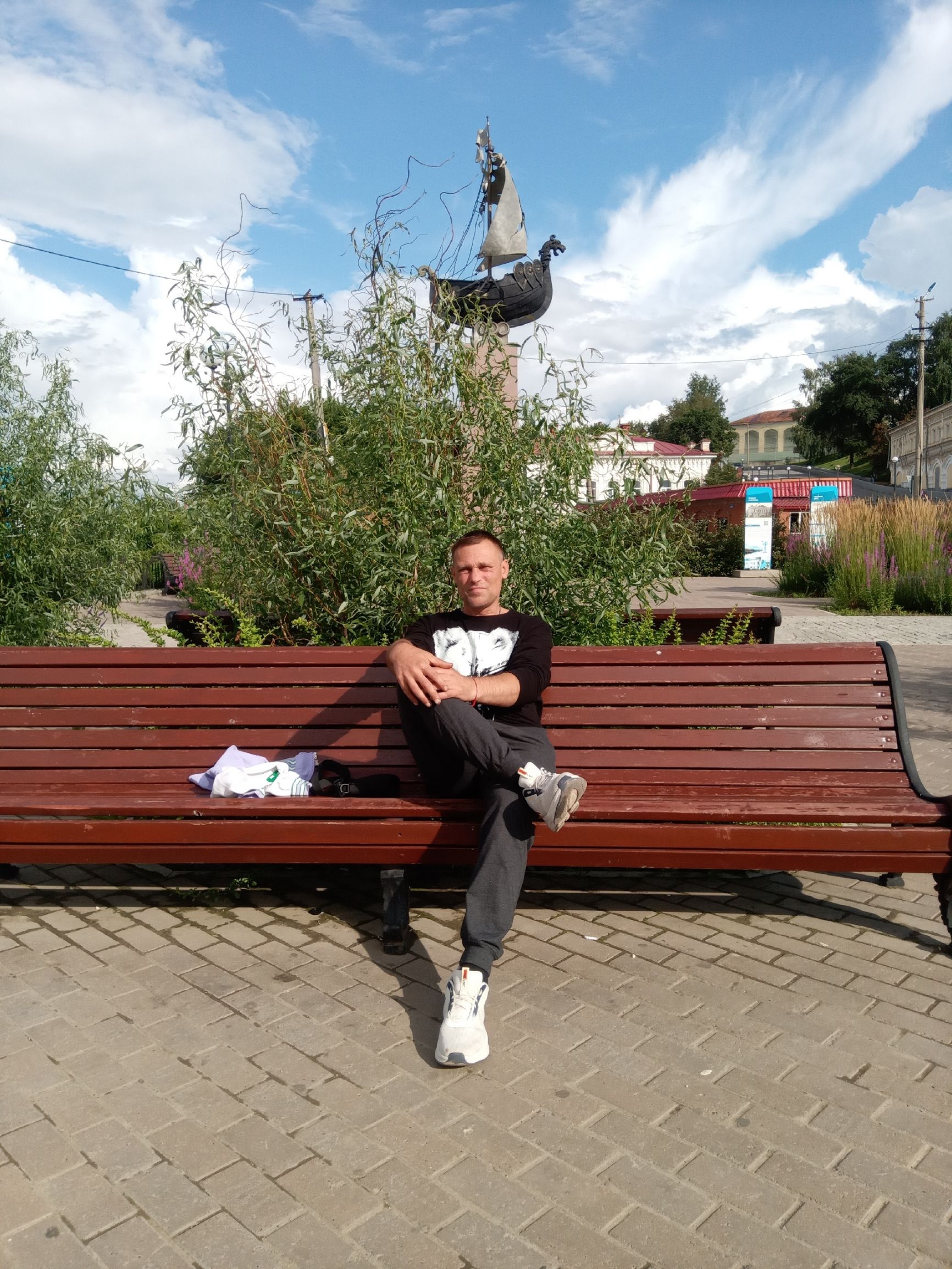 Мужчина сидит на деревянной скамейке в парке; на фоне кусты и небо.