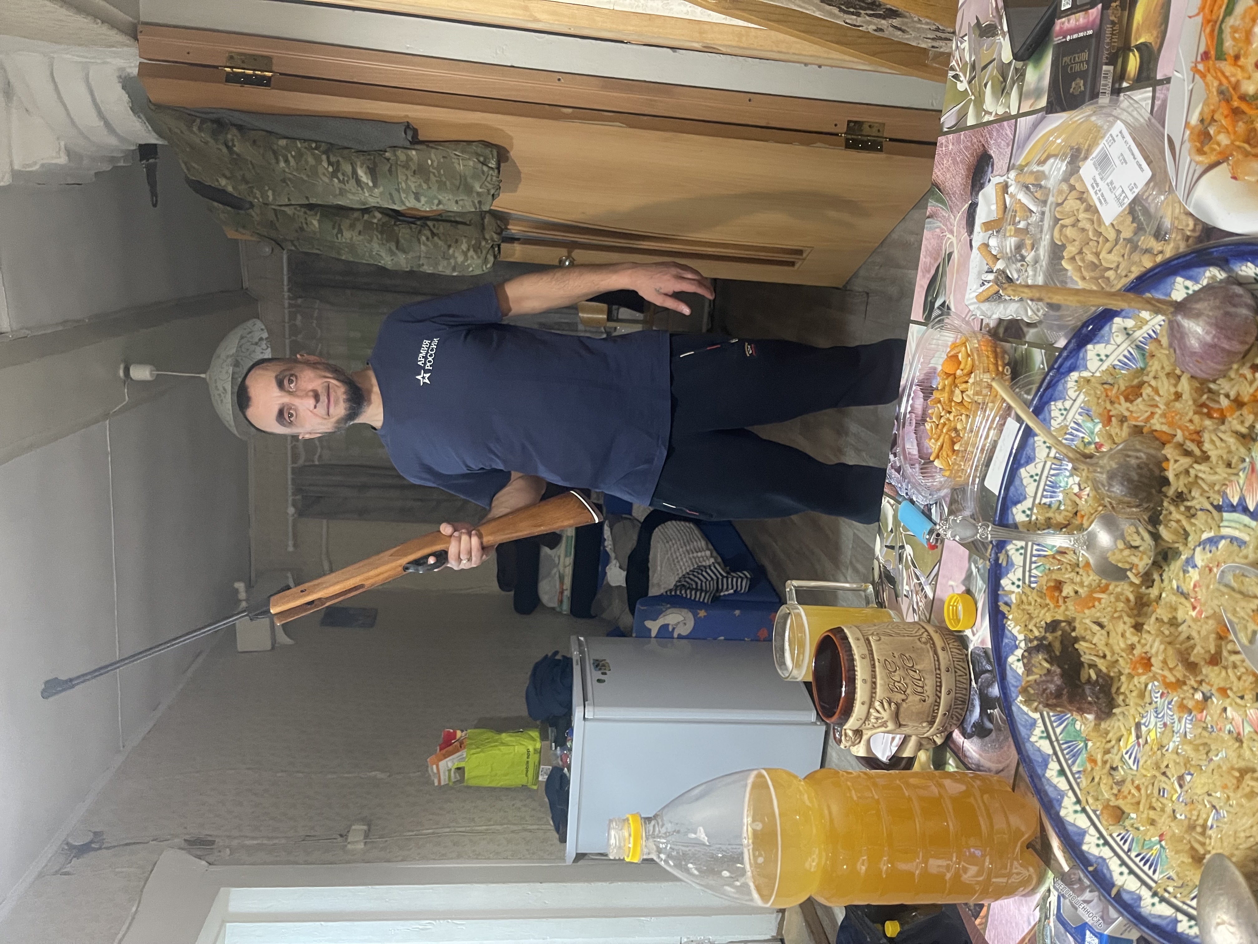 Человек в кухне держит длинное оружие (ружье) на фоне стола с едой.