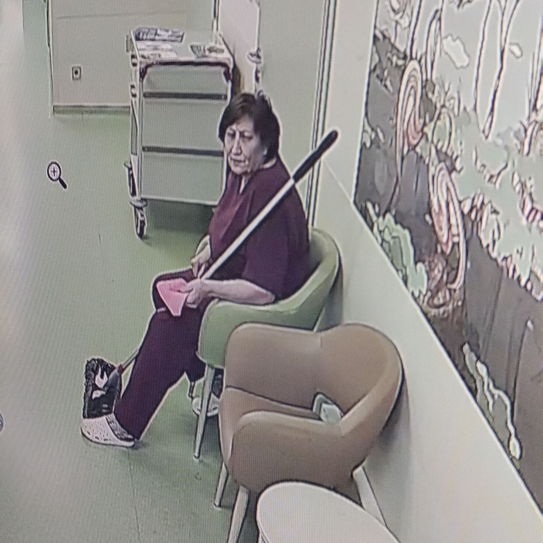 Женщина сидит в помещении на кресле, рядом стул и рисунки на стене, в руках длинный предмет (похоже на палку/венчик).