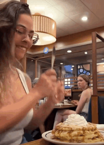 Женщина в кафе улыбается и режет вилкой большой десерт с взбитыми сливками