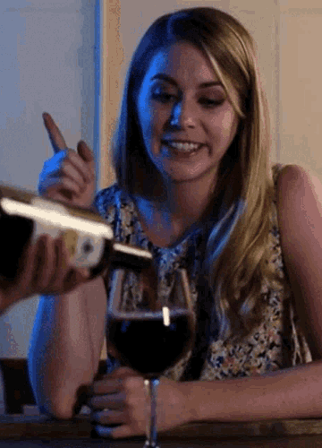 Женщина наливает вино за столом, улыбается и поднимает палец.