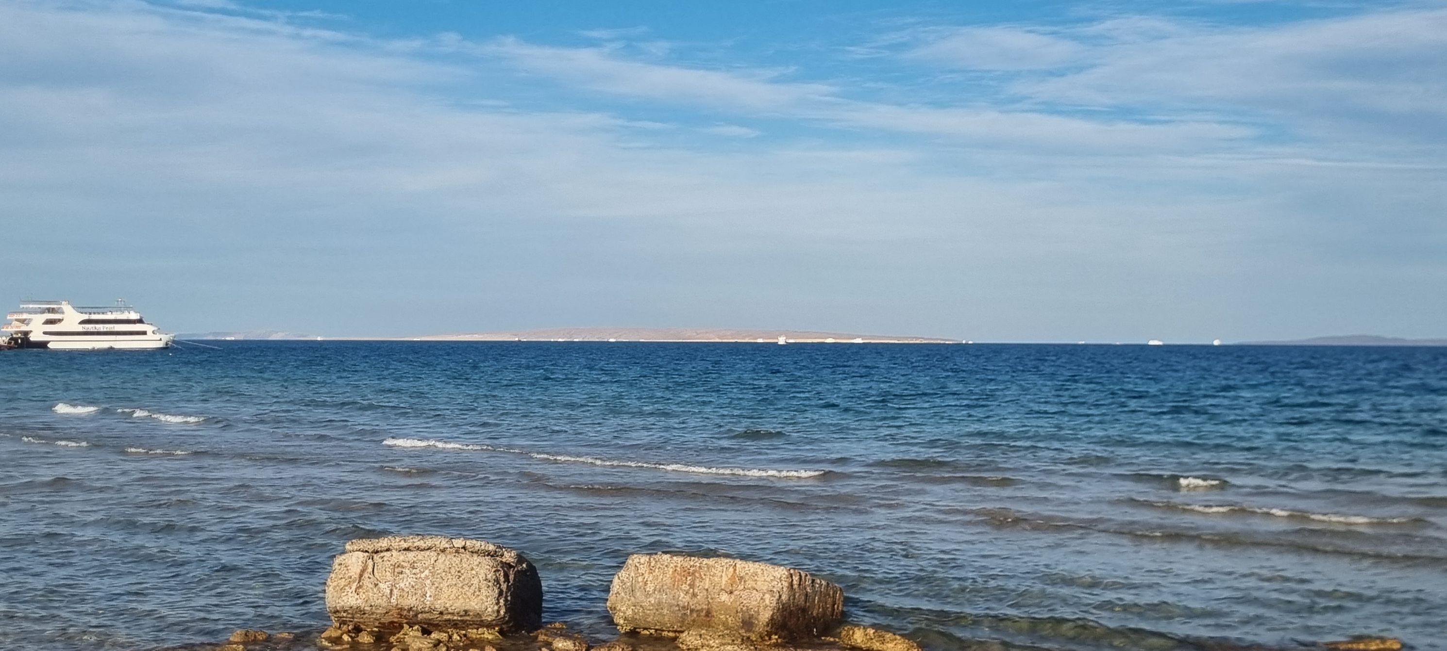 морской берег с камнями на переднем плане и яхтой вдалеке