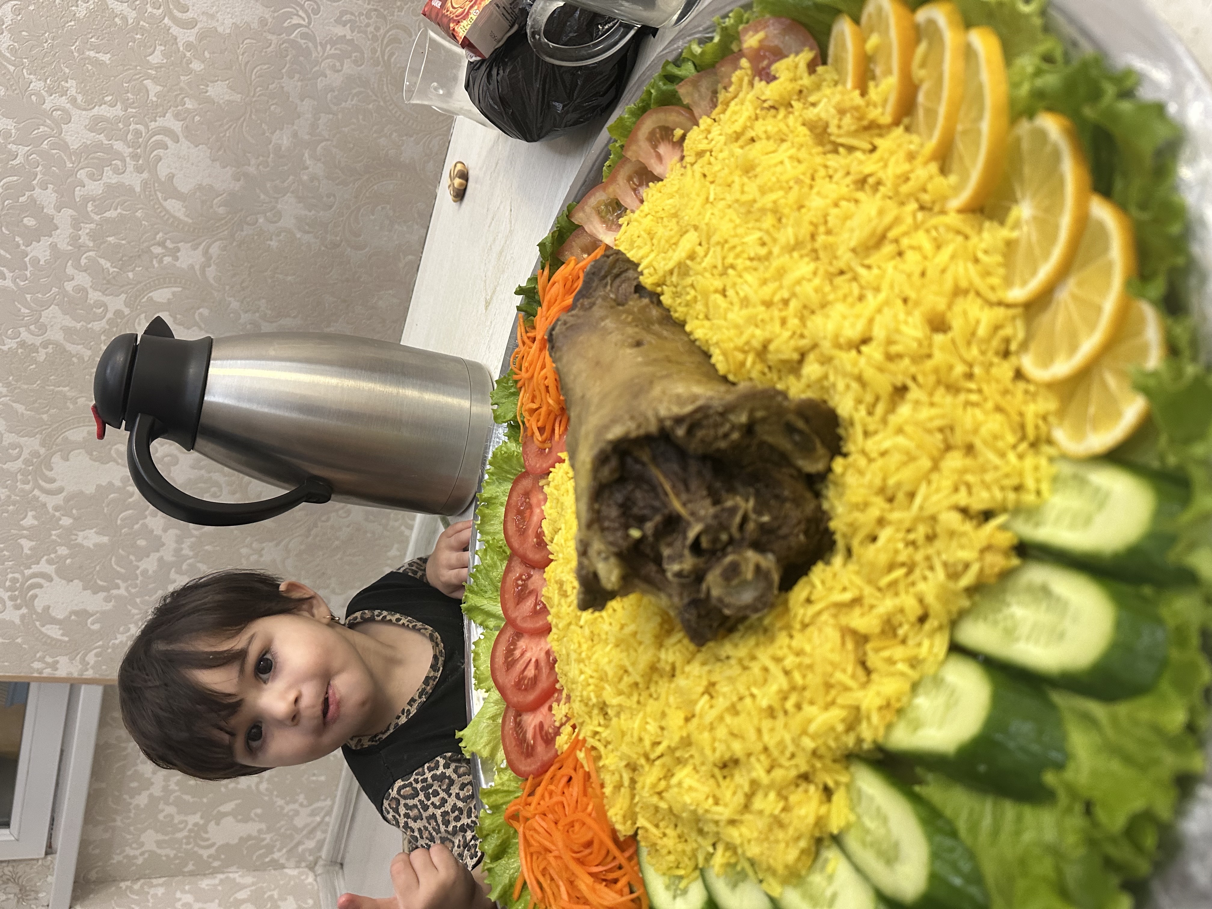 Человек стоит возле большого блюда с рисом и овощами на тарелке, на середине — темная часть блюда, возможно мясо животного.