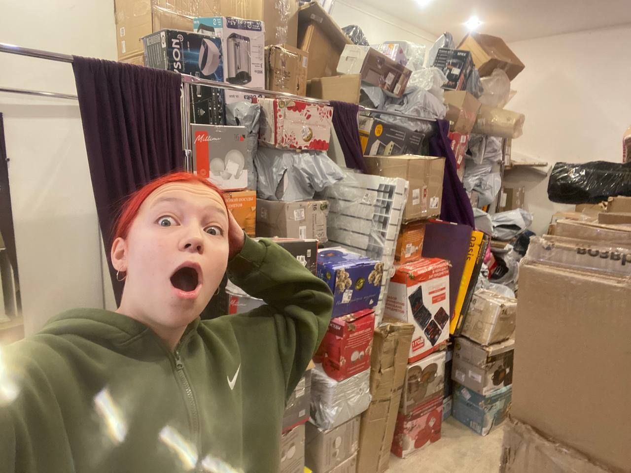 Человек делает селфи в складском помещении возле стопки коробок и упаковок.