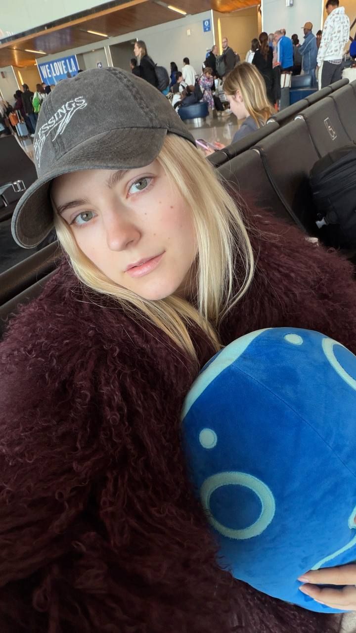 Девушка в аэропорту сидит с синей подушкой и шарфом/курткой бордового цвета