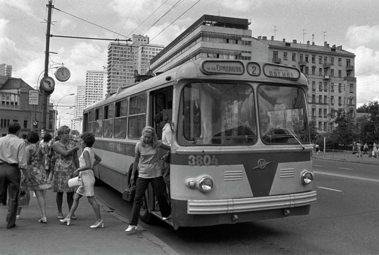 Черно-белое фото старого трамвая/автобуса на улице, люди вокруг, дети выходят из транспорта.