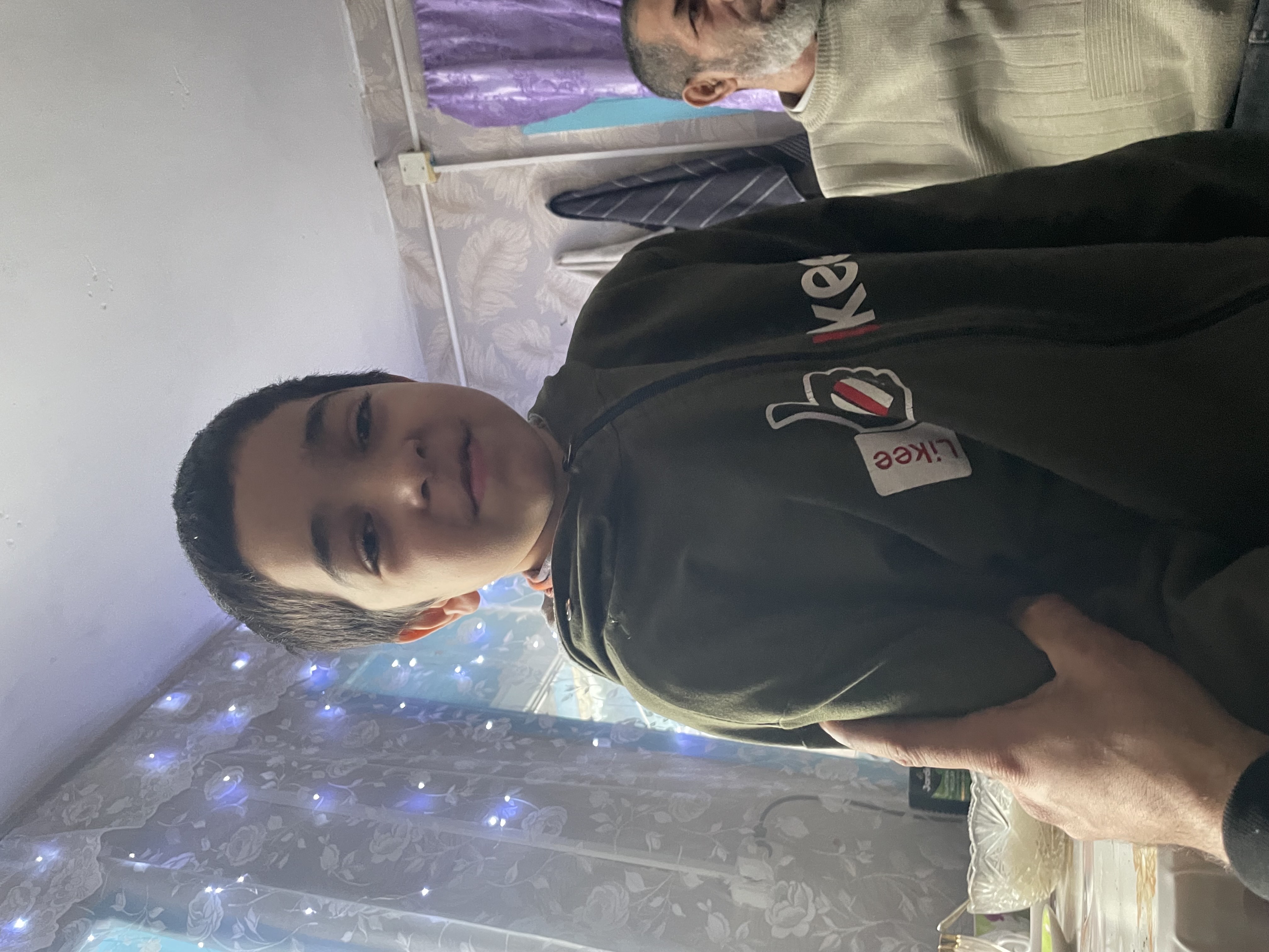 Молодой мужчина в зелёной толстовке pose в помещении, фоном ещё человек и штора.