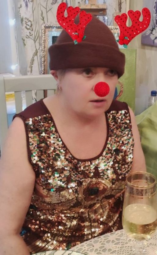 Женщина в блестящей кофте с рождественскими атрибутами: красный нос, ушки-олени, шапка и бокал у стола.