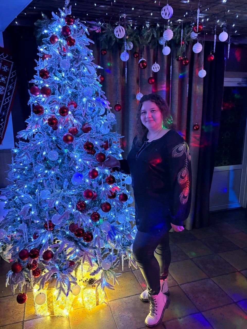 Женщина стоит возле белой ёлки с голубыми огнями и красными шарами на празднике.