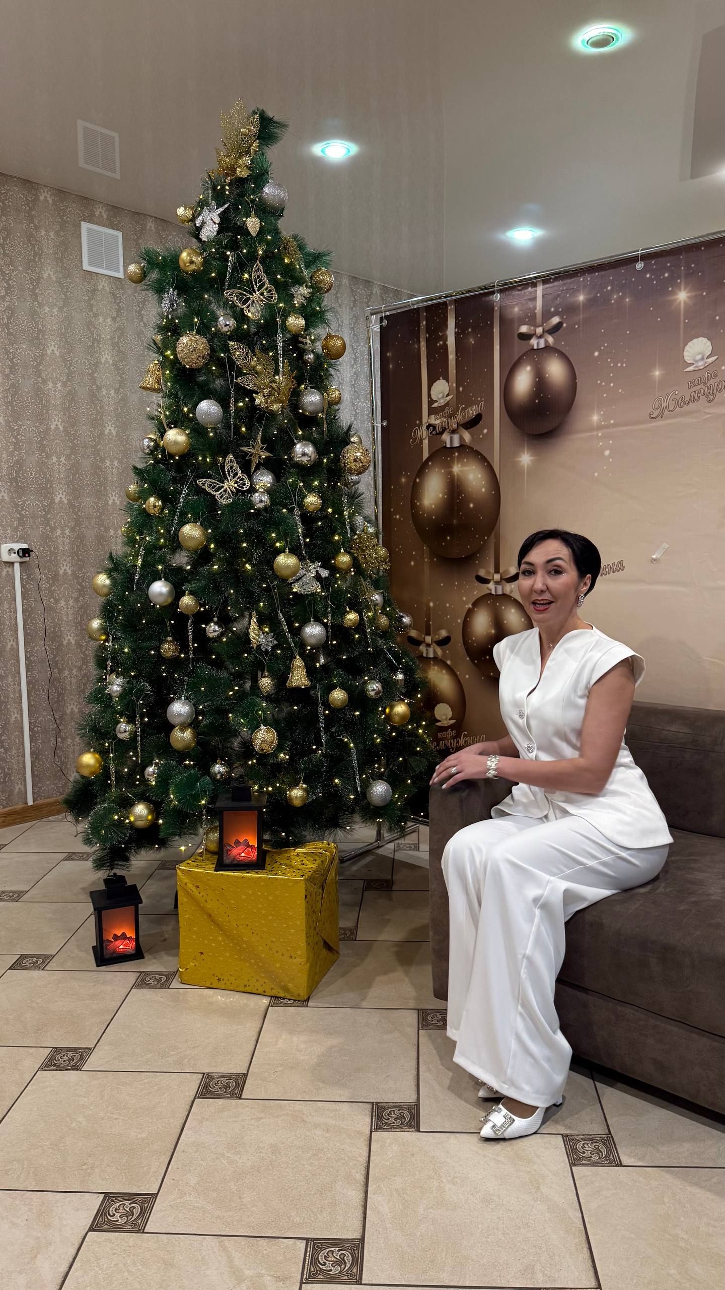 Женщина в белом сидит рядом с украшенной рождественской ёлкой в уютной комнате.
