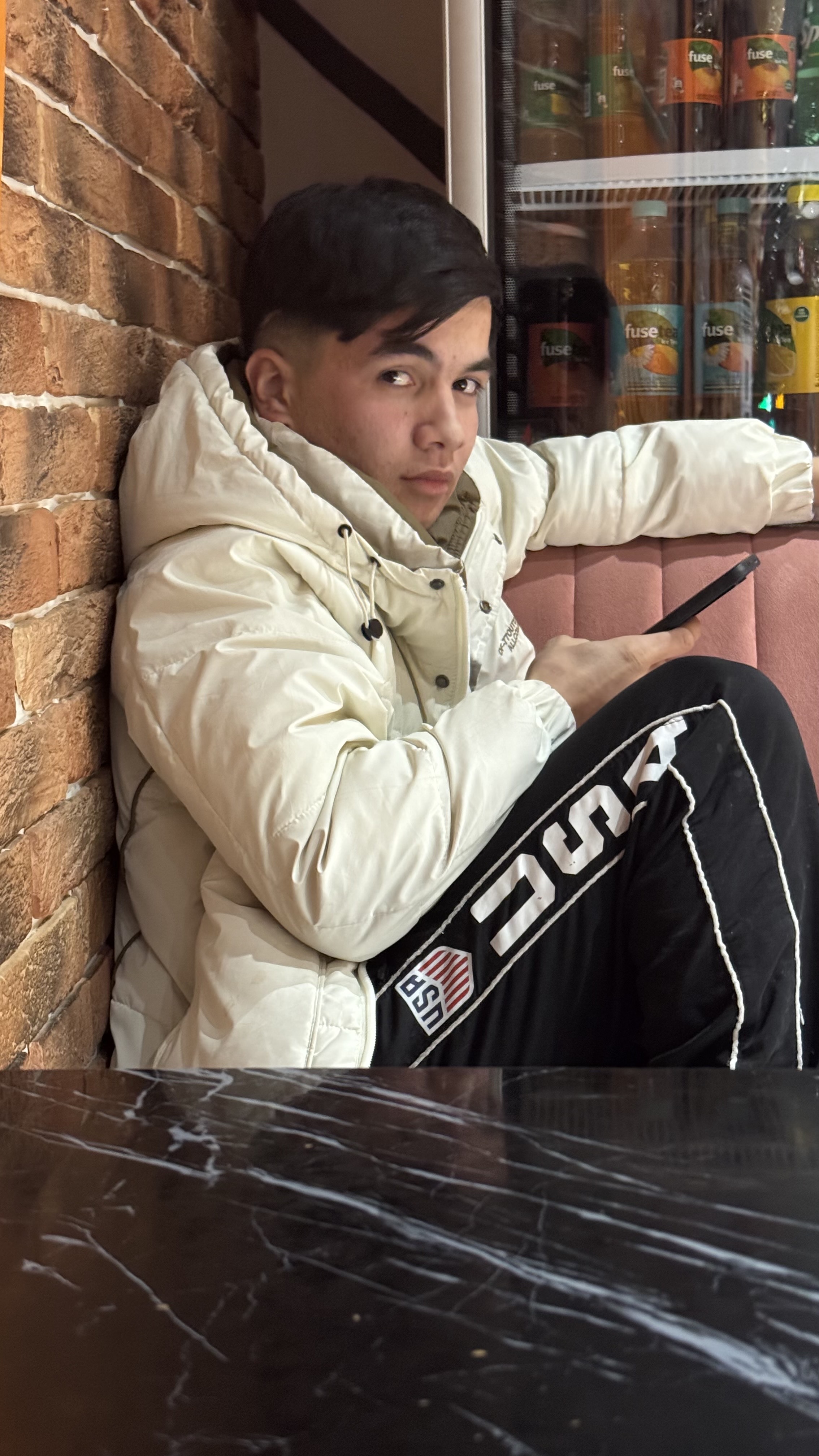 Парень в белой куртке сидит у кирпичной стены в кафе и держит телефон.