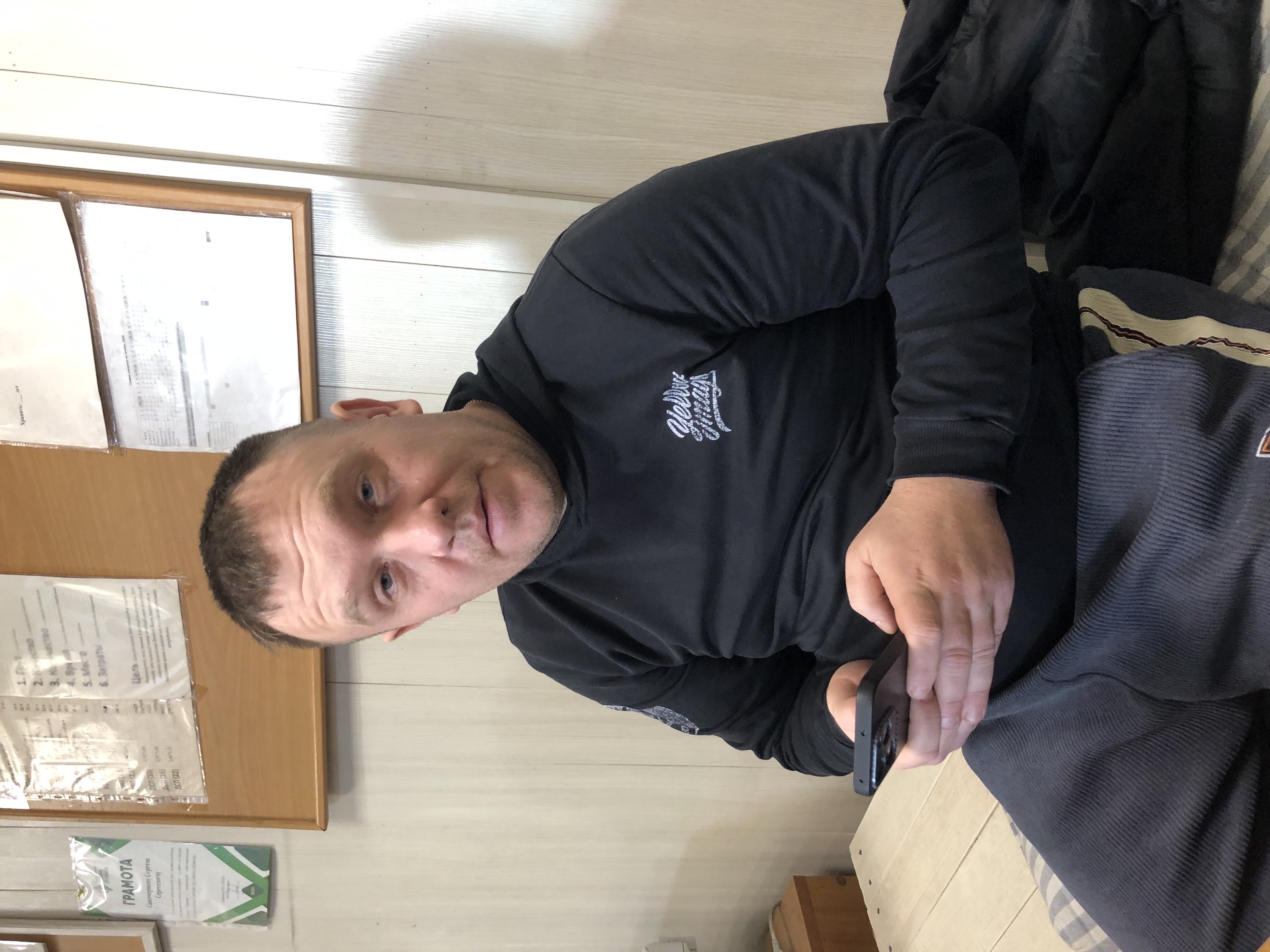 Мужчина в черной одежде сидит на стуле в помещении, держит телефон.