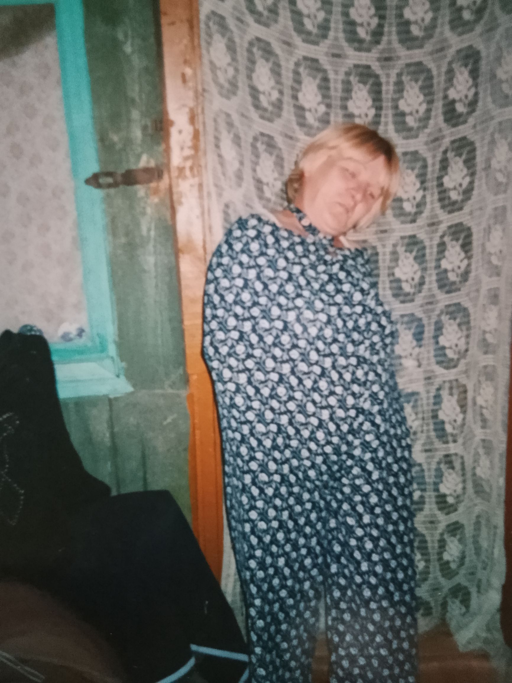 Пожилая женщина в длинном платье стоит в комнате с узорной занавеской.