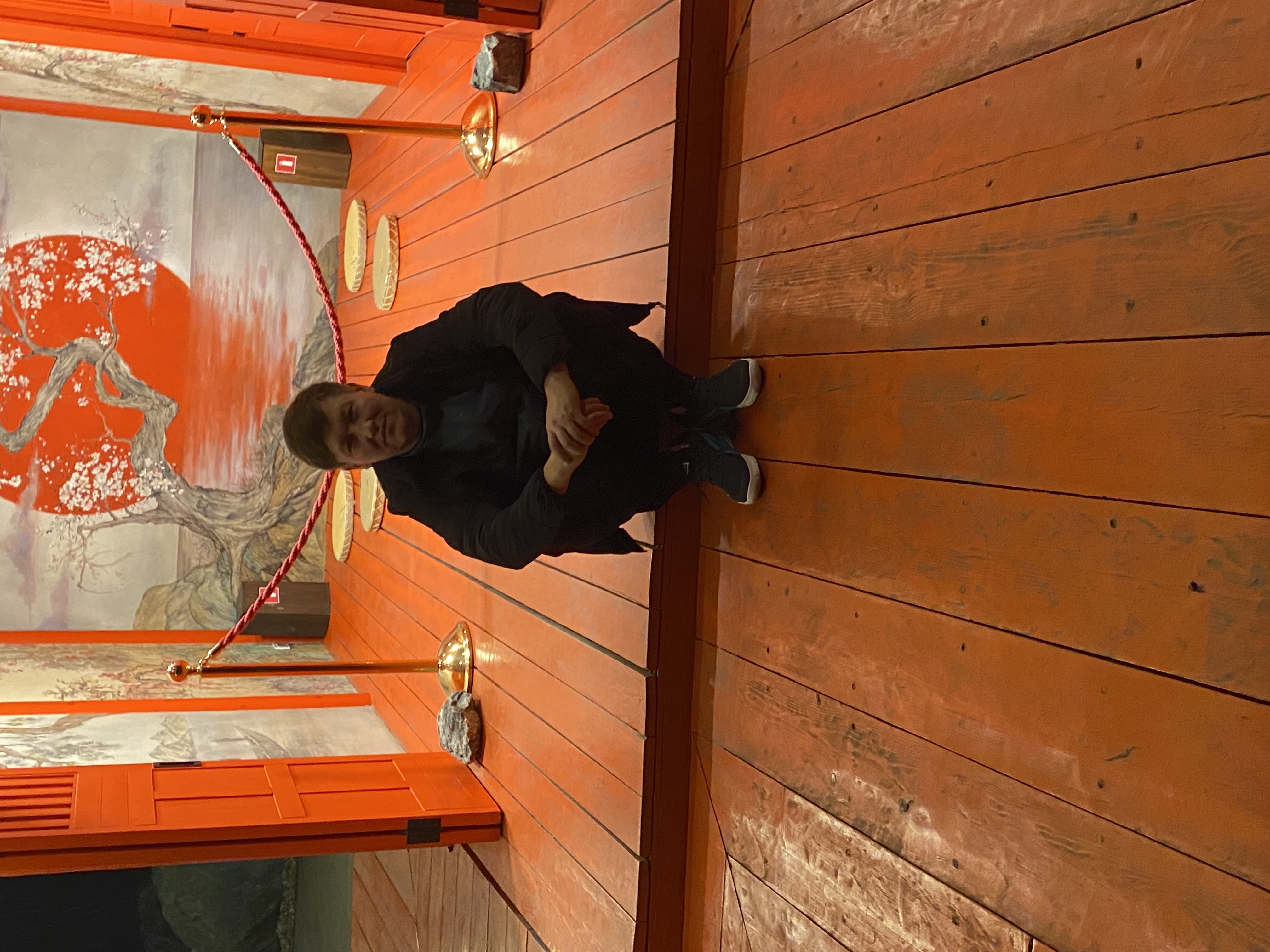 Человек стоит/сидит в тёплом деревянном помещении спортивной зоны с оборудованием на стенах