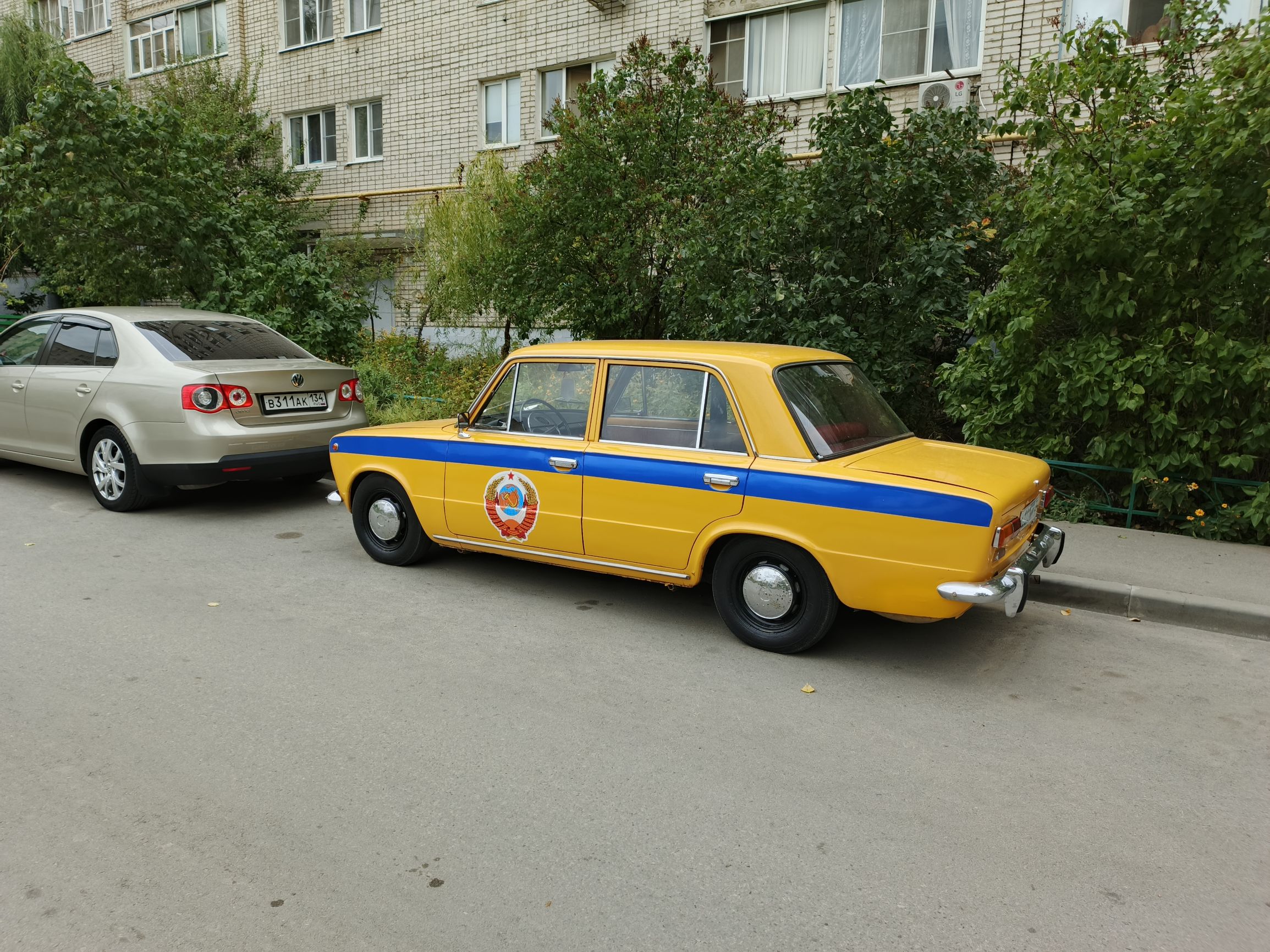 Жёлтый ретро-автомобиль с синей полосой на парковке возле многоэтажки