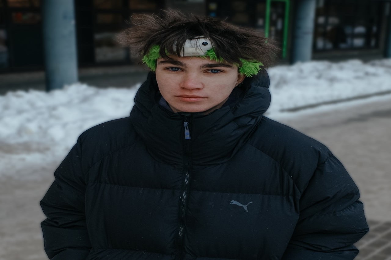 Молодой человек в пуховике на улице, на голове гирлянда из зелени