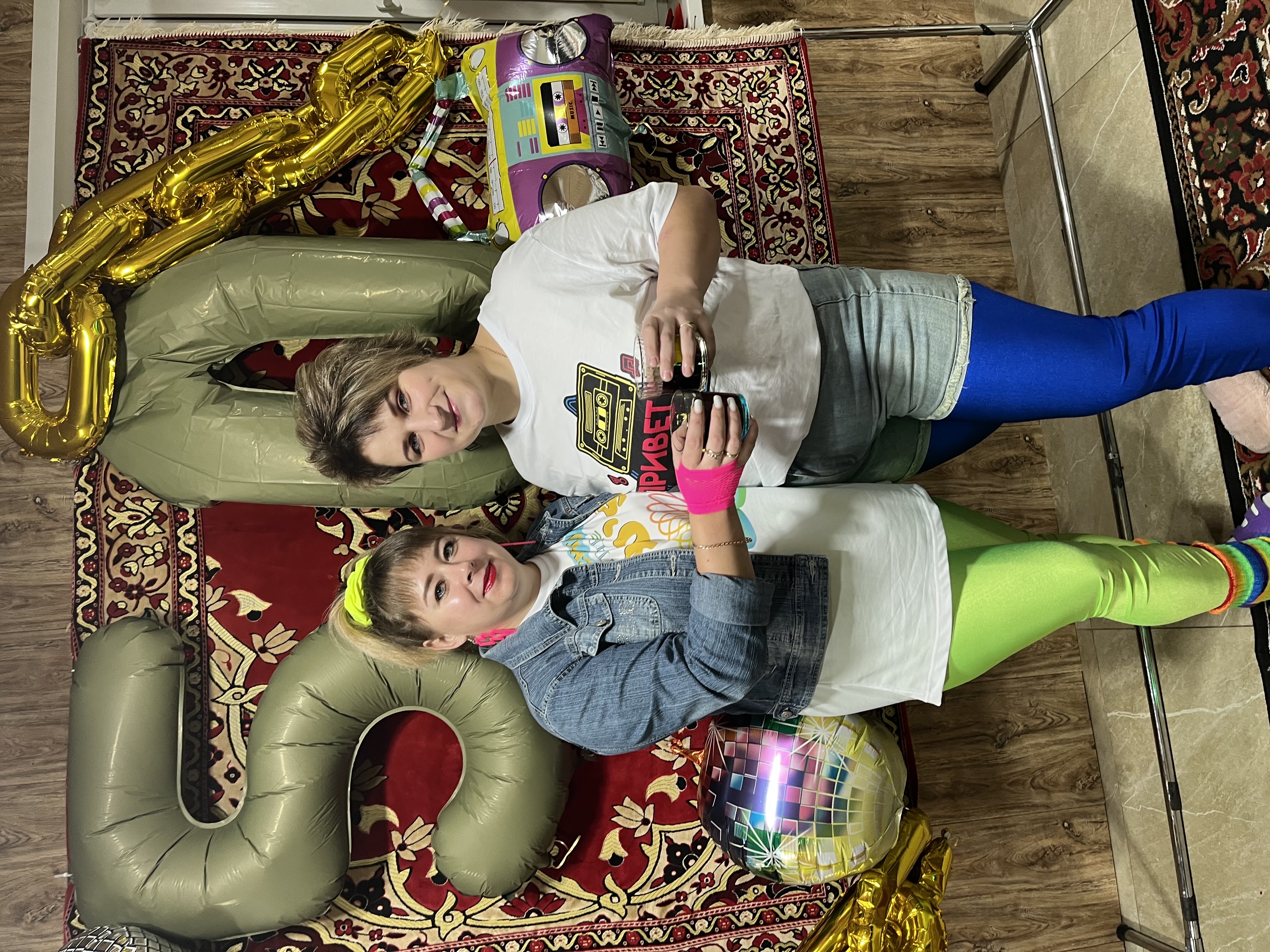 На фото две девушки держат подарочную карту/сюрприз, позируют рядом с большими золотыми шариками и буквой «2» на ковре.