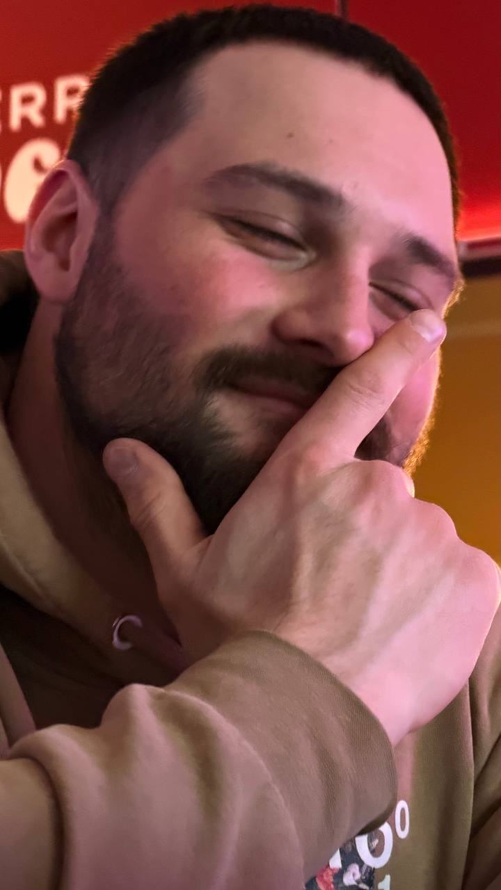 Мужчина улыбается, приподняв руку к лицу и касаясь носа/губ.