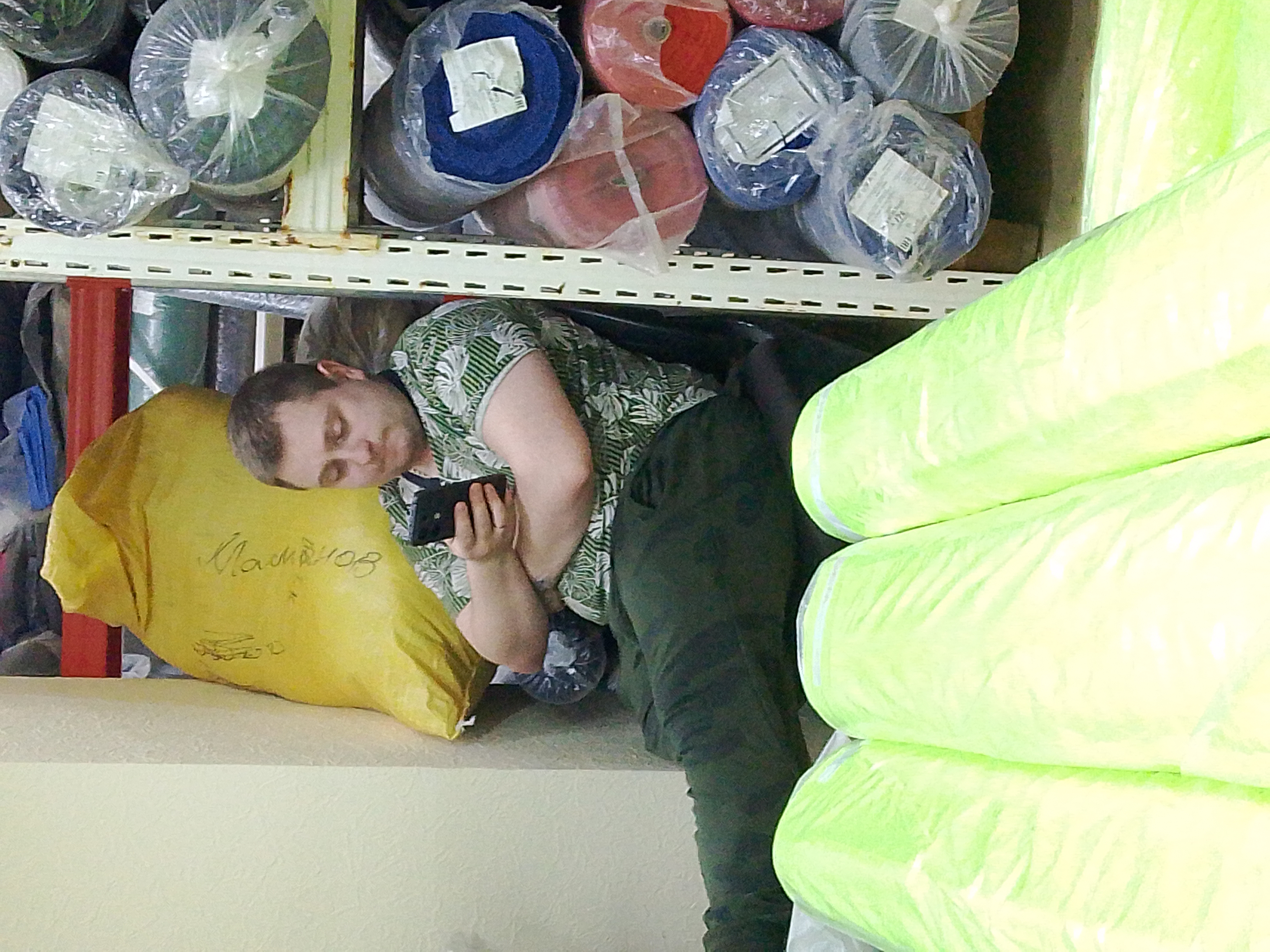 Человек сидит на полу склада среди мешков и рулонов ткани.