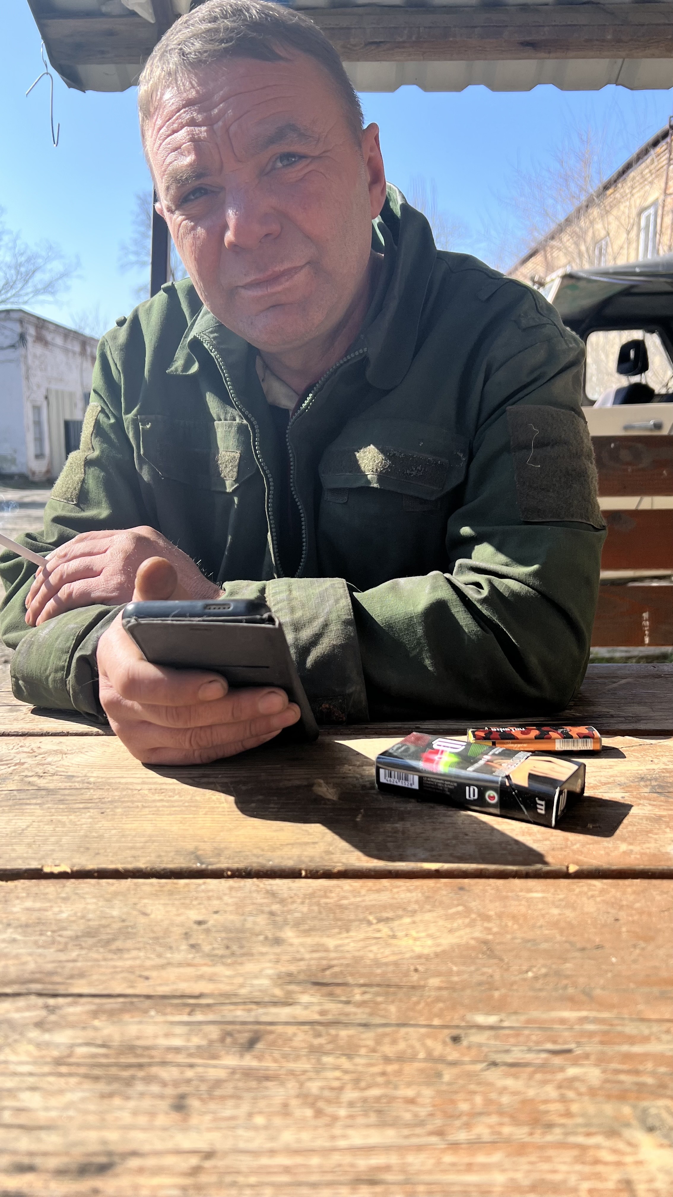 Мужчина за столиком на улице держит телефон, рядом пачки сигарет на столе.