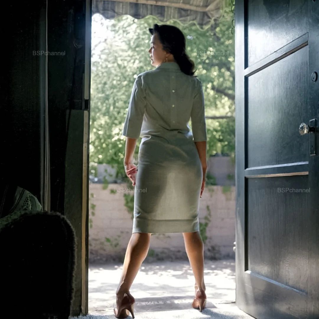 Женщина в светлом платье стоит на пороге открытой двери, вид сзади.