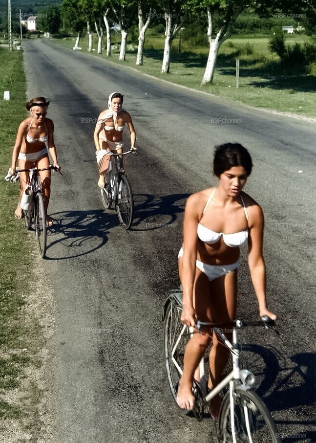 Три женщины в купальниках едут на велосипедах по сельской дороге.