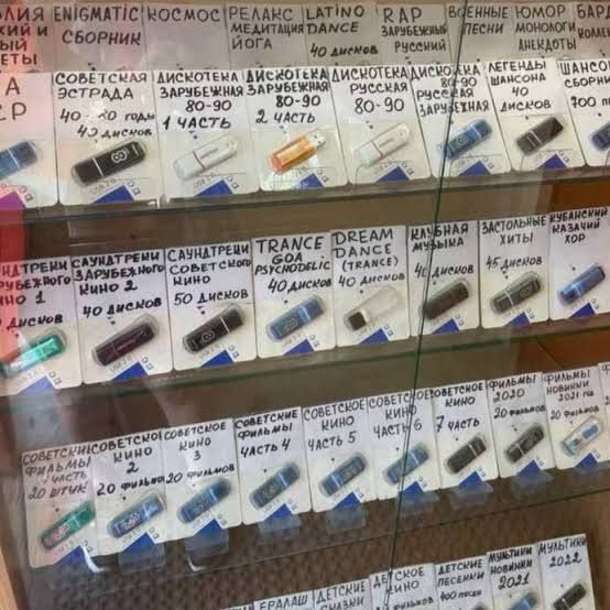 На витрине представлены USB-флешки с различными категориями медиаконтента. Видны надписи: ENIGMATIC, КОСМОС, РЕЛАКС, LATINO, RAP, ВОЕННЫЕ ЮМОР, БАРД, и сборник, МЕДИТАЦИЯ DANCE, ЙОГА, 40 дисков, ЗАРУБЕЖНЫЙ, РУССКИЙ, ПЕСНИ, МОНОЛОГИ, АНЕКДОТЫ, 700 песен. Также есть категории: СОВЕТСКАЯ ЭСТРАДА, ДИСКОТЕКА ЗАРУБЕЖНАЯ 80-90 (1 и 2 часть), ДИСКОТЕКА