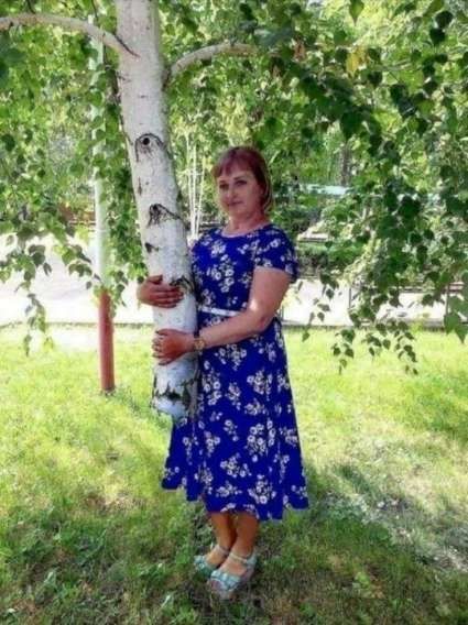 Женщина в синем платье с цветочным принтом стоит и обнимает белое дерево в парке.