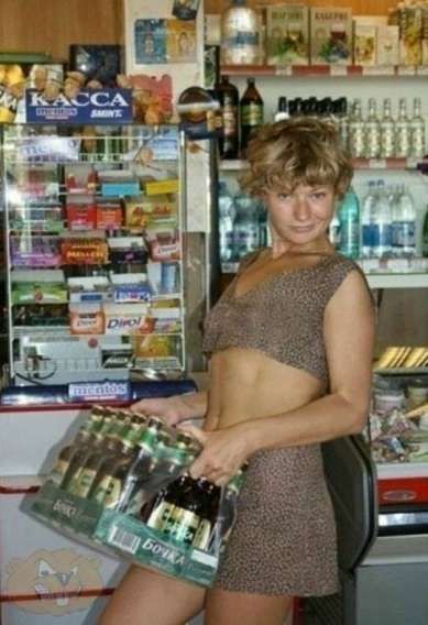 Женщина в леопардовом платье позирует в продуктовом магазине у витрины с напитками, держит поднос с бутылками.