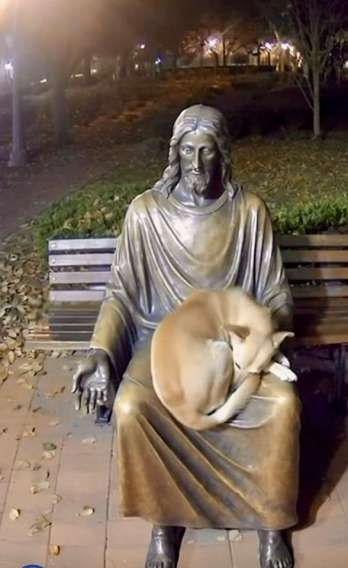 Статуя Иисуса на скамейке, собака лежит на коленях.