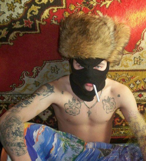 Мужчина без футболки в балаклаве и ушанке, курит сигарету на фоне ковра.