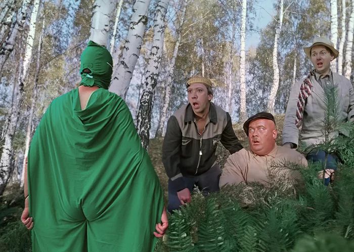 Кадр в лесу: три мужчины в камуфляже/лесной одежде, один в ярко-зеленом костюме, создаётся ощущение комедийной сцены.