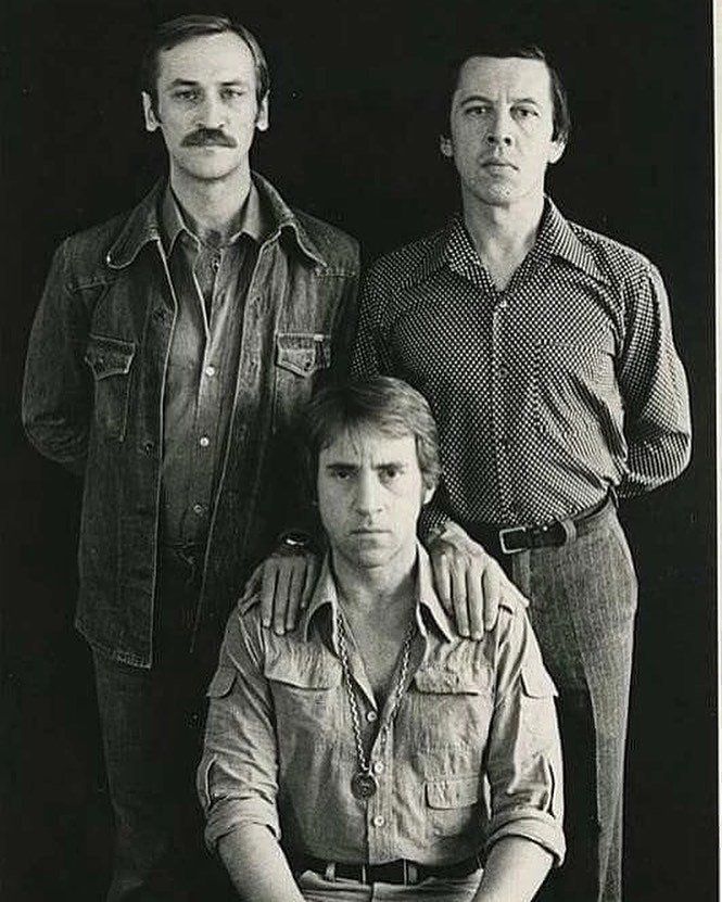 Черно-белая фото трёх мужчин: один сидит спереди, двое стоят позади и держат руки на его плечах.