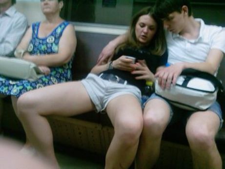 Люди на метро, слева женщина в ярком платье, справа пара сидит близко, парень держит сумку на коленях, девушка смотрит в телефон.