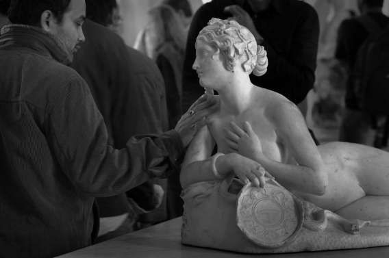 Человек трогает обнаженную статую женщины на столе в людной обстановке.