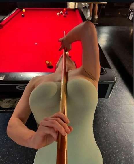 Девушка в облегающем платье делает фото над пул-соусом, держась за кий с позиции над головой.