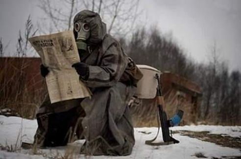 Человек в противогазе читает газету на снегу, рядом оружие