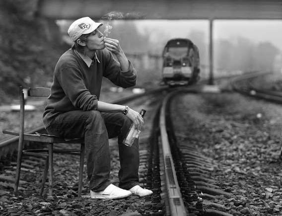Мужчина сидит на стуле на железнодорожной колее и держит бутылку, к нему приближаётся поезд.