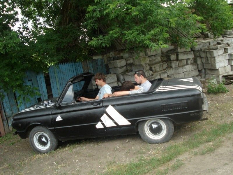 Два мужчины в черном кабриолете старого образца на проселке возле стены.