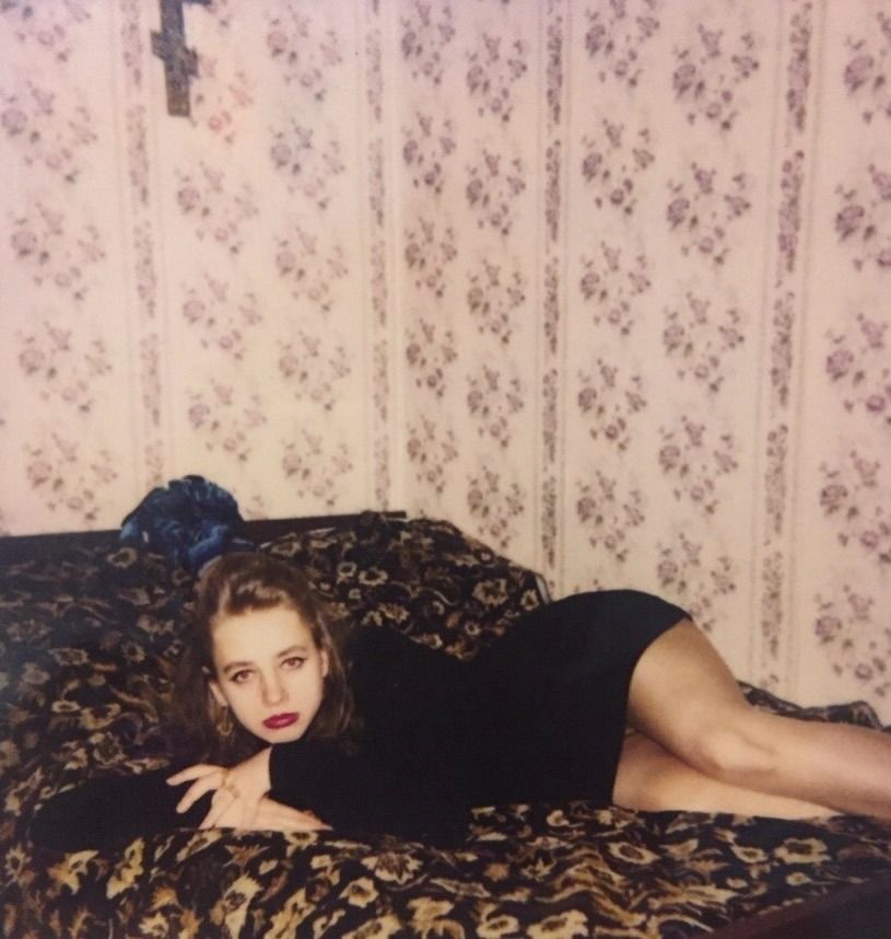 Девушка в черном платье лежит на кровати рядом с узорным покрывалом.