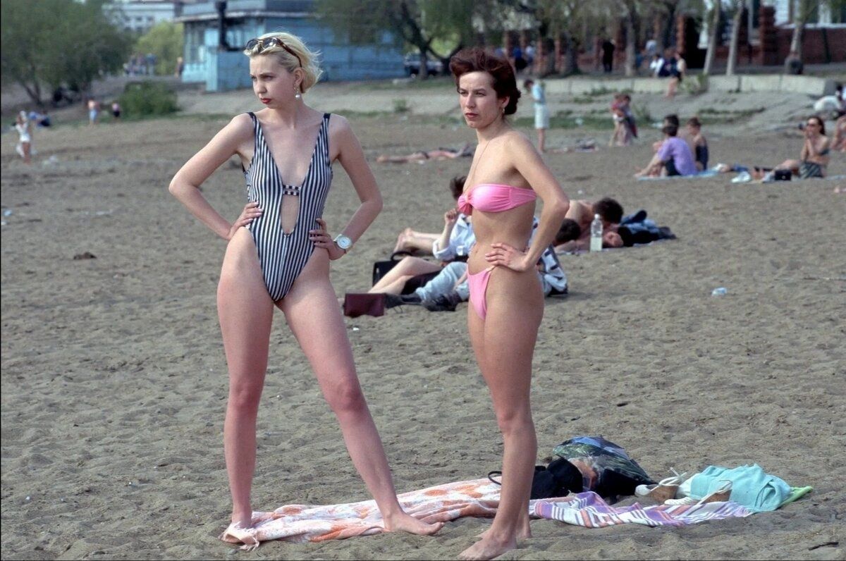 Две девушки в купальниках стоят на пляже, за ними люди на песке.