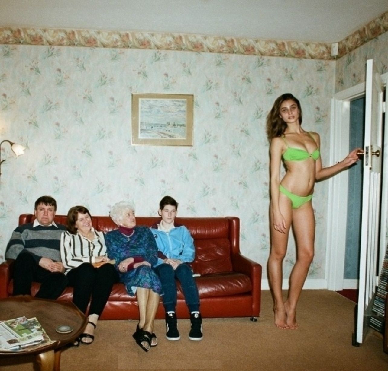Семейная гостиная: за диваном сидят взрослые люди, молодая женщина в зелёном бикини стоит у двери.
