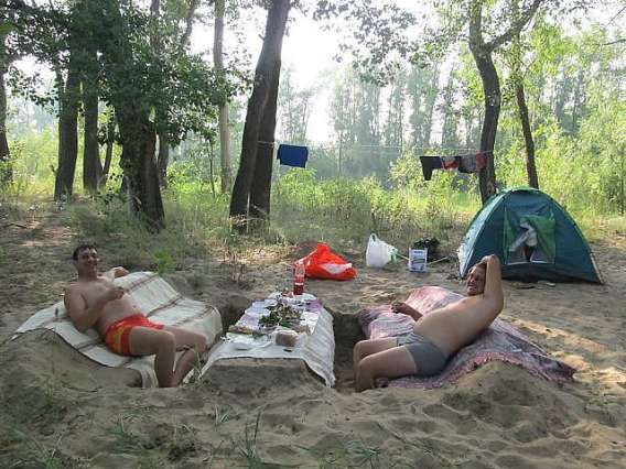 Два взрослых человека на походном кемпинге в лесу, рядом палатка и стол с едой.