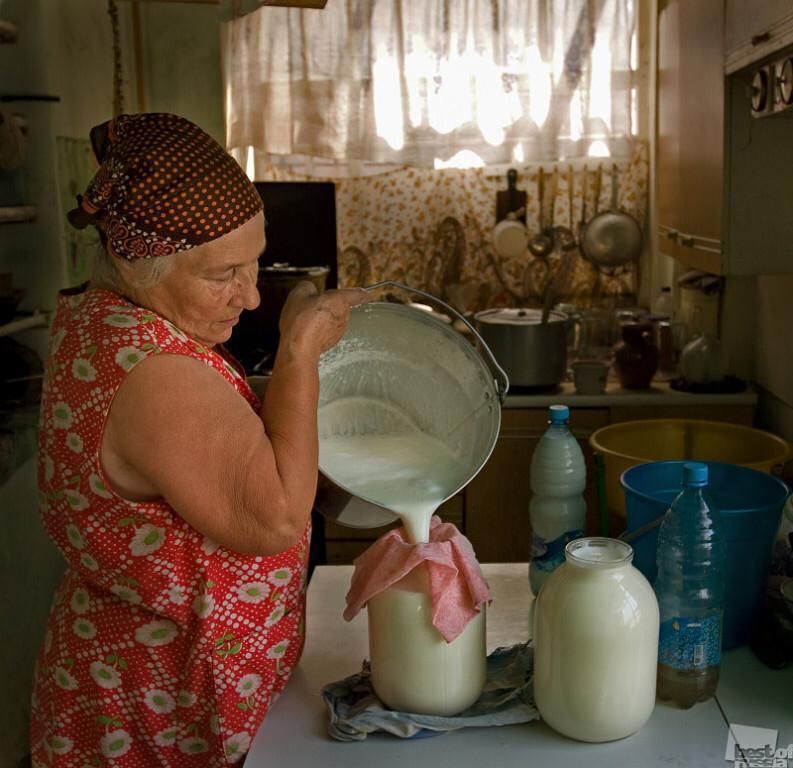 Старушка в кухне наливает молоко в бутылки из большого кувшина