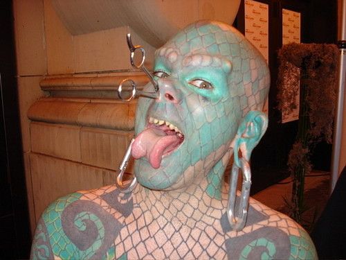 Человек с синей сетчатой раскраской лица и тела, пирсингами и языком наружу.