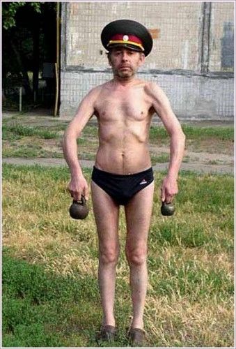 Мужчина в военном кепи стоит на улице в шортах без одежды, держит гирьки