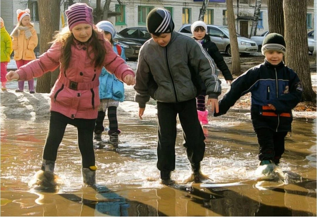 Группа детей играет в лужах во дворе на прогулке.