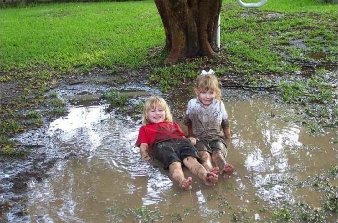 Два ребенка сидят в луже на траве, радостно улыбаются.