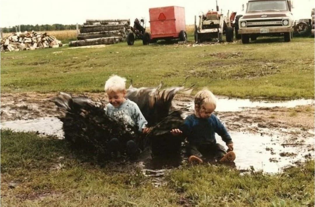 Два ребенка сидят в грязной луже, рядом расправляют крылья крупные птицы