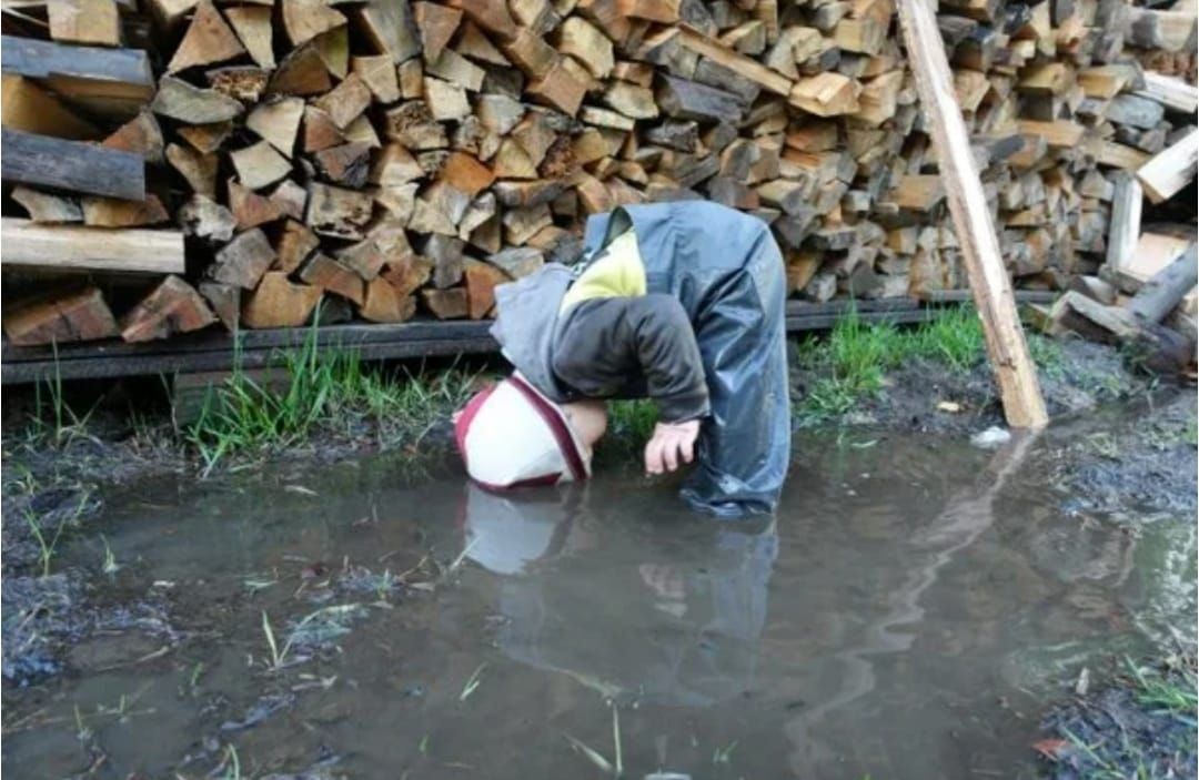 Человек в грязной воде возле склада дров, нагнувшись лицом в воду.
